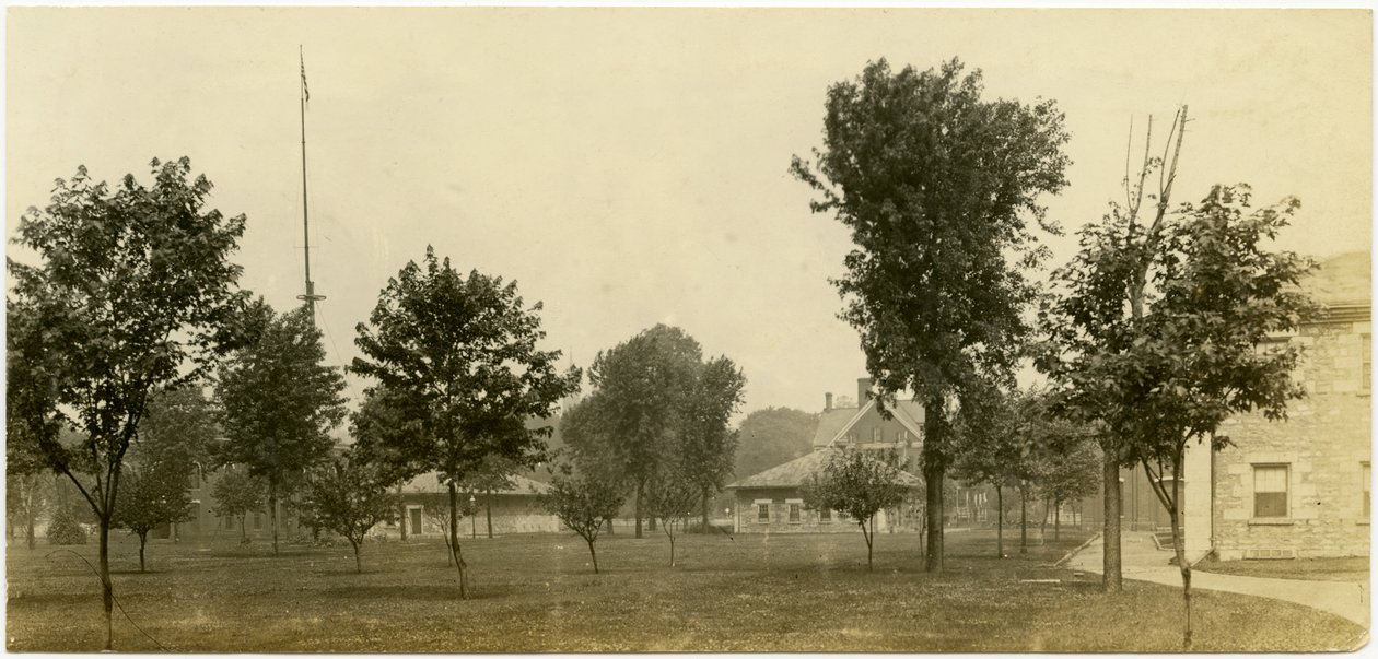 Horizontale, sepia foto van een groep bakstenen gebouwen grenzend aan een open groene ruimte met verspreide bomen. Sommige bakstenen gebouwen lijken huizen te zijn door Carl Michel