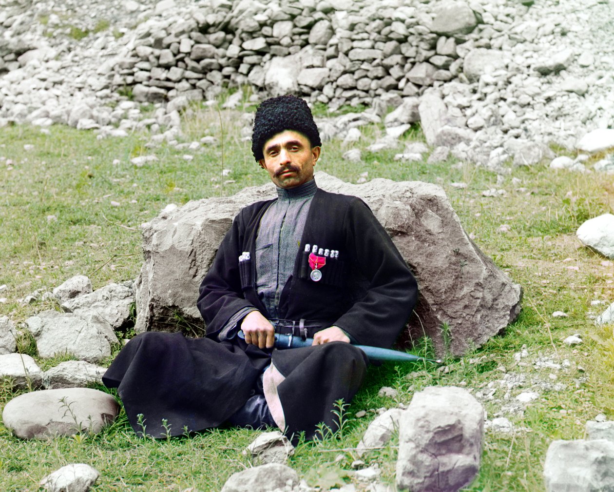 Dagestaanse types - Soennitische moslimman in traditionele kleding en hoofddeksel, met een geschede dolk aan zijn zijde, zittend op de grond, Russische Rijk, tussen 1905 en 1915 door Sergey Prokudin Gorsky